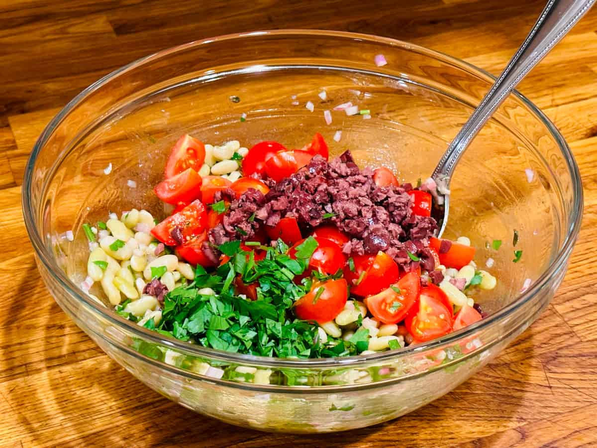 Flageolet beans, quartered cherry tomatoes, chopped parsley, and chopped olives in a glass bowl with a steel spoon.