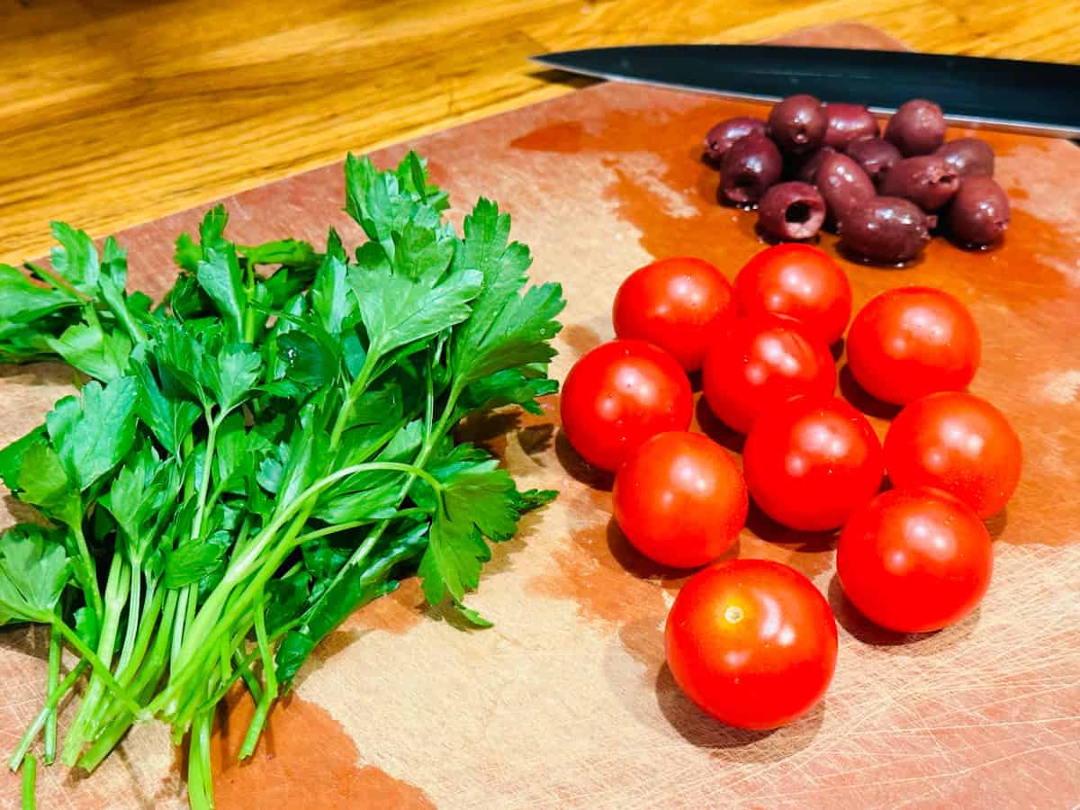 Several sprigs of parsley, some cherry tomatoes, and a pile of small black olives on a cutting board in front of a knife.
