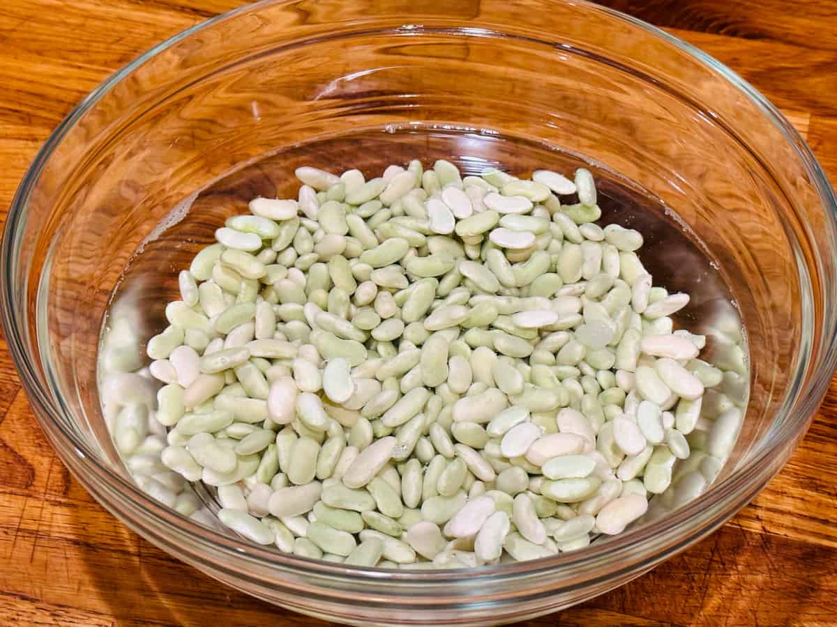 Dried pale green beans soaking in water in a large glass bowl.