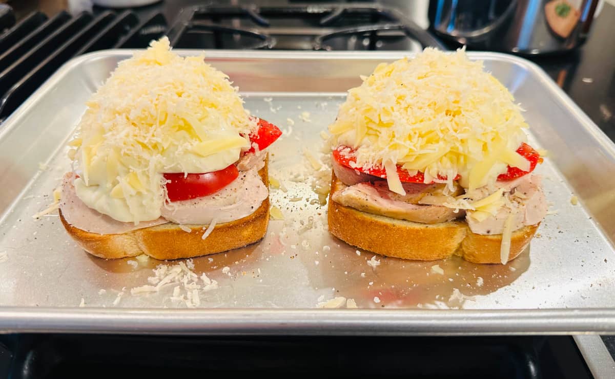 Two partially assembled open faced sandwiches with roasted turkey and tomatoes covered in shredded cheese on a metal baking sheet.