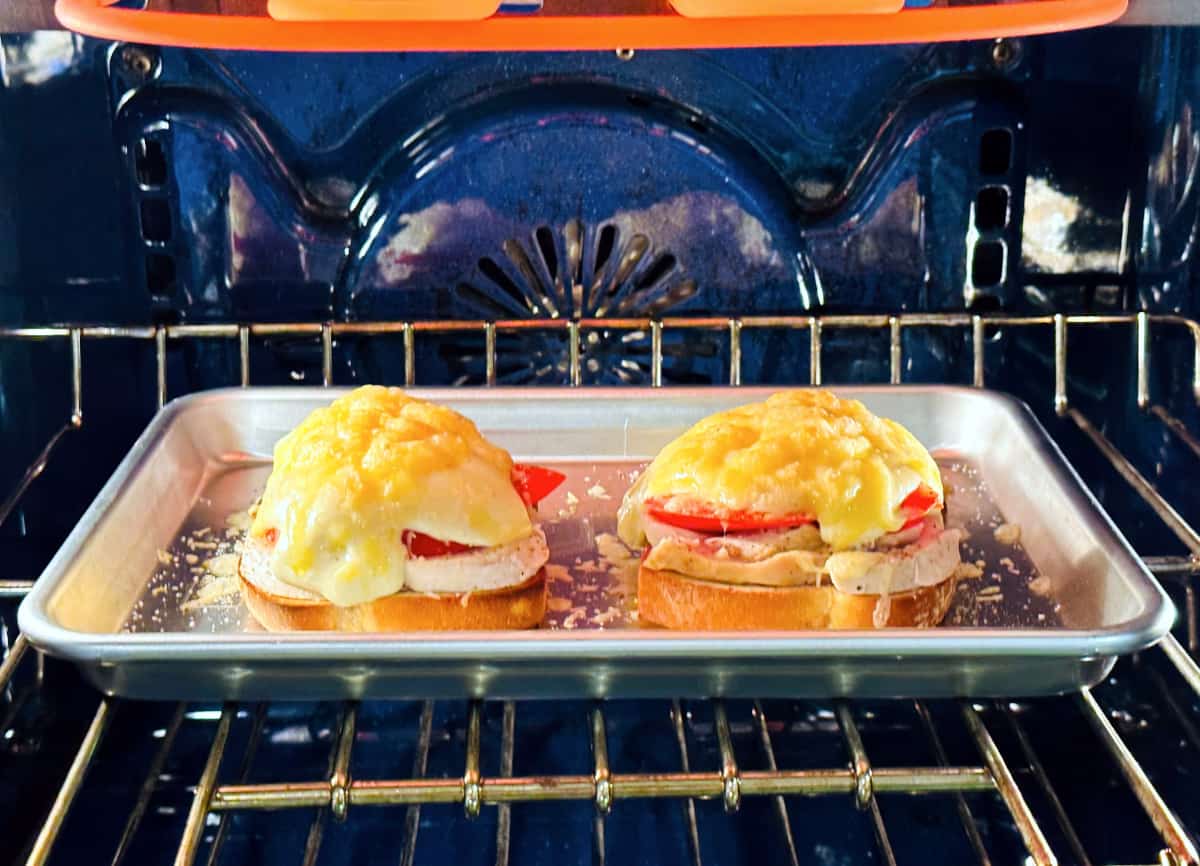 Two open faced sandwiches with roasted turkey and tomatoes covered in cheese sauce on a metal baking sheet toasting under a broiler.