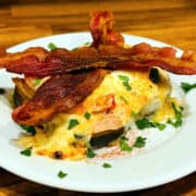 Open faced sandwich with roasted turkey and tomatoes covered in cheese sauce, sprinkled with parsley and paprika, and topped with two slices of bacon sitting on a white plate.