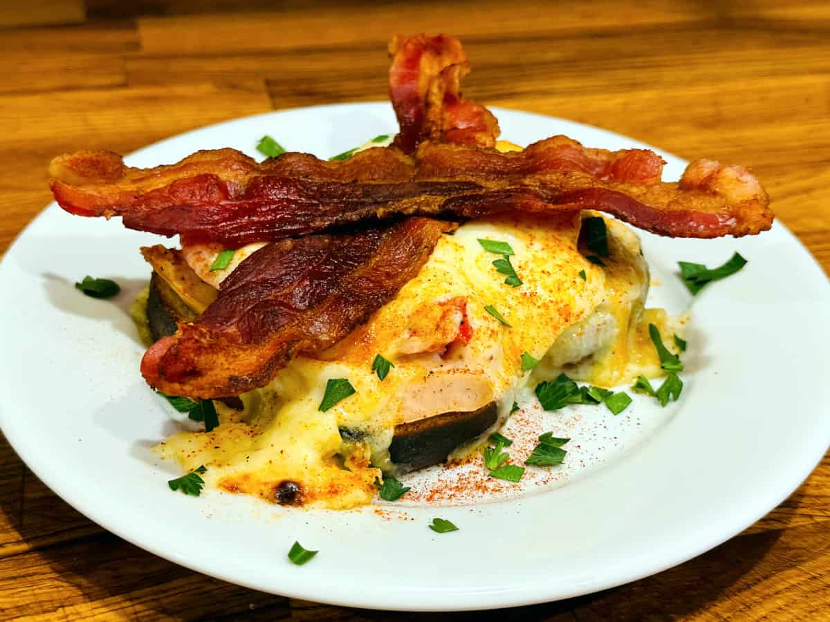 Open faced sandwich with roasted turkey and tomatoes covered in cheese sauce, sprinkled with parsley and paprika, and topped with two slices of bacon sitting on a white plate.