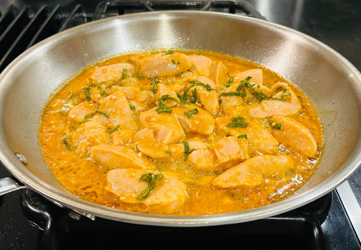 Chicken in a golden brown liquid with chopped mint cooking in a large steel saucepan.