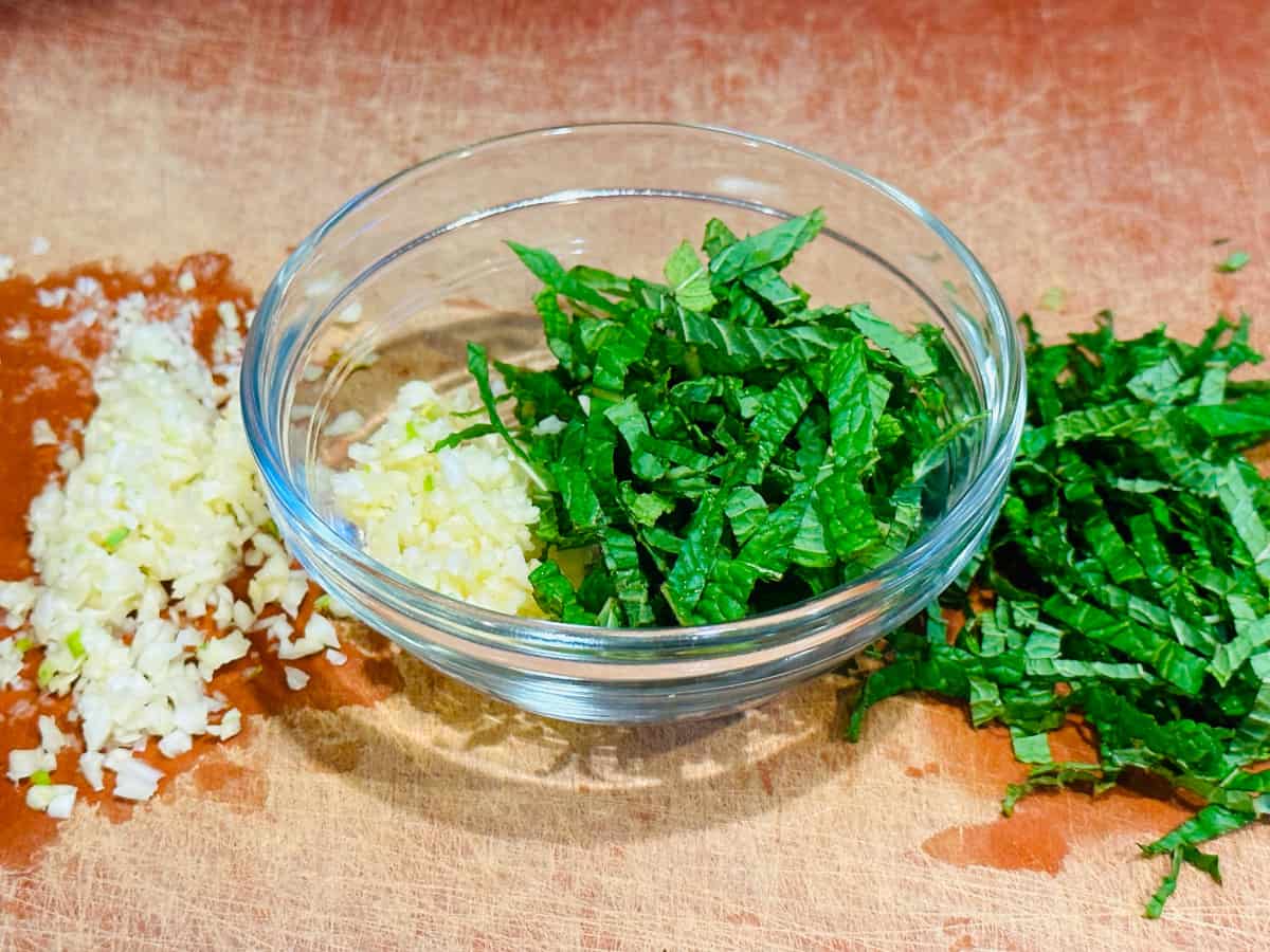 Minced garlic and finely chopped mint in a glass bowl next to garlic on one side and mint on the other.
