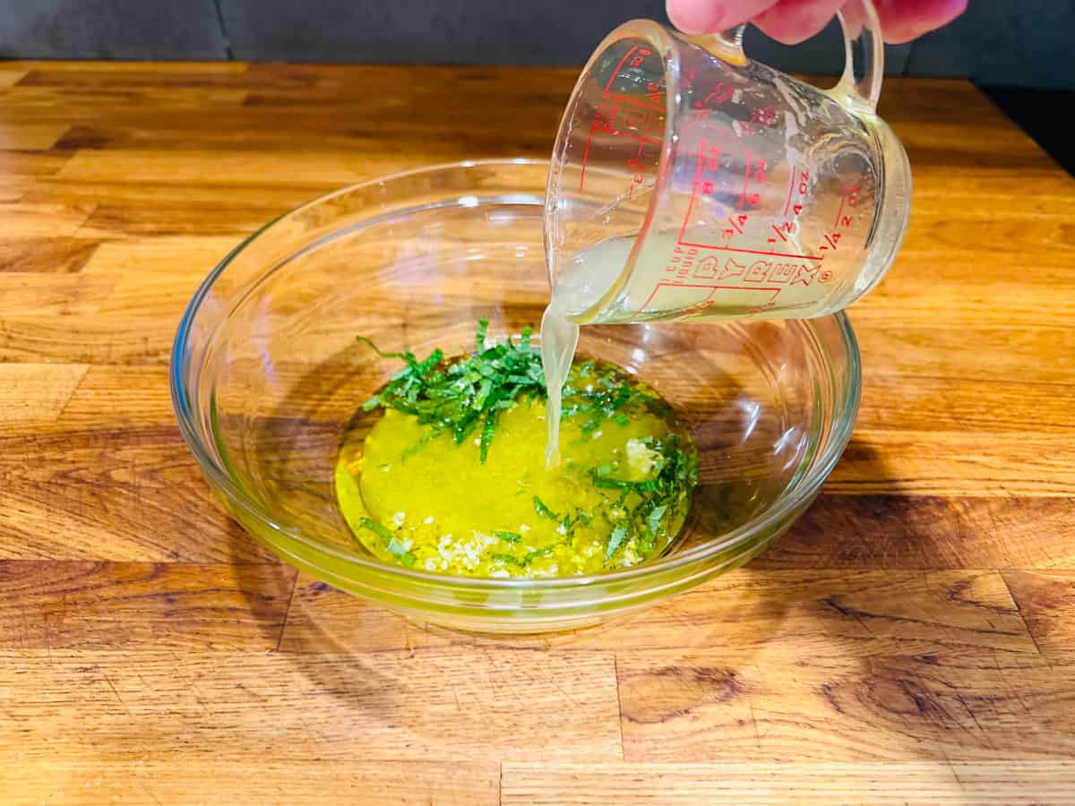 Lemon juice being poured from a glass measuring cup into a glass bowl containing chopped garlic and mint.