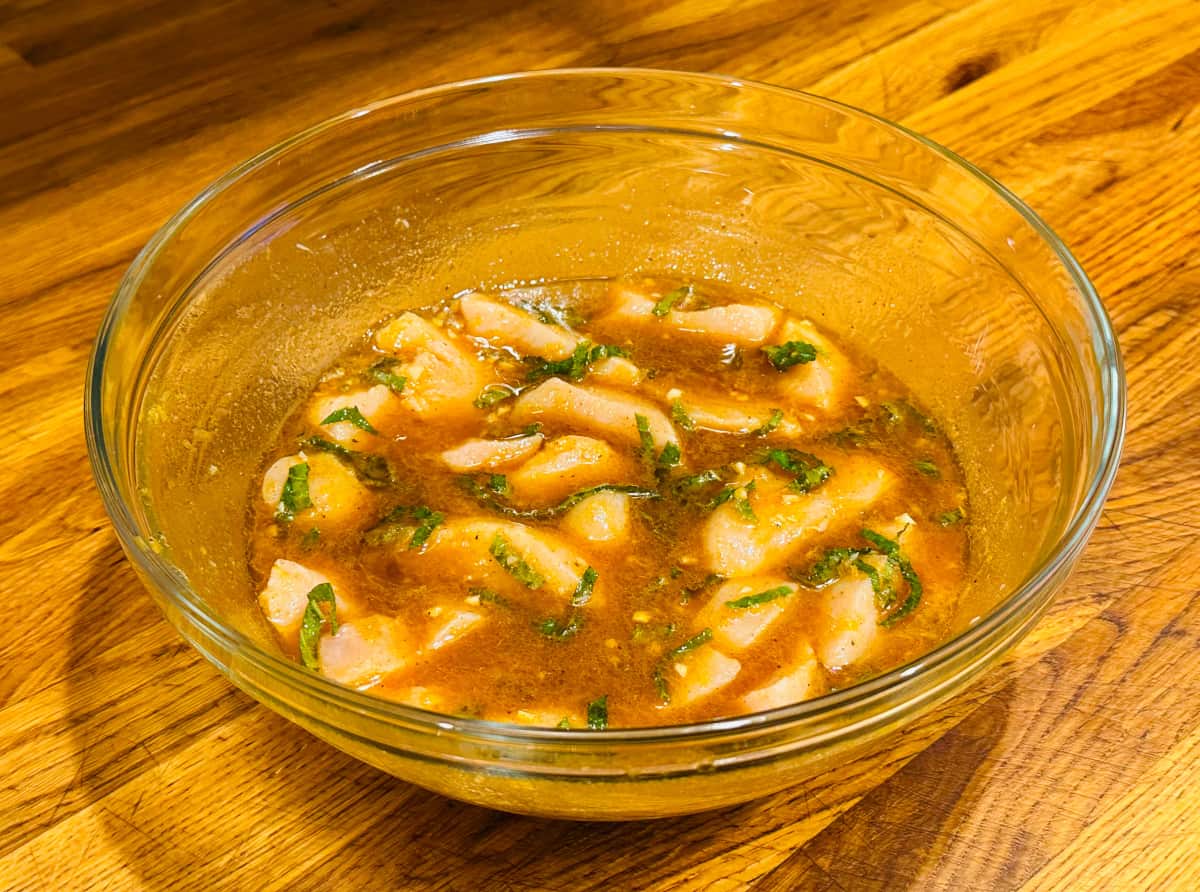 Raw chunks of chicken marinading in brown liquid with flecks of chopped mint in a glass bowl.