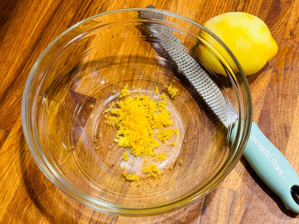 Lemon zest in a glass bowl next to a microplane with a light blue handle and a zested lemon.