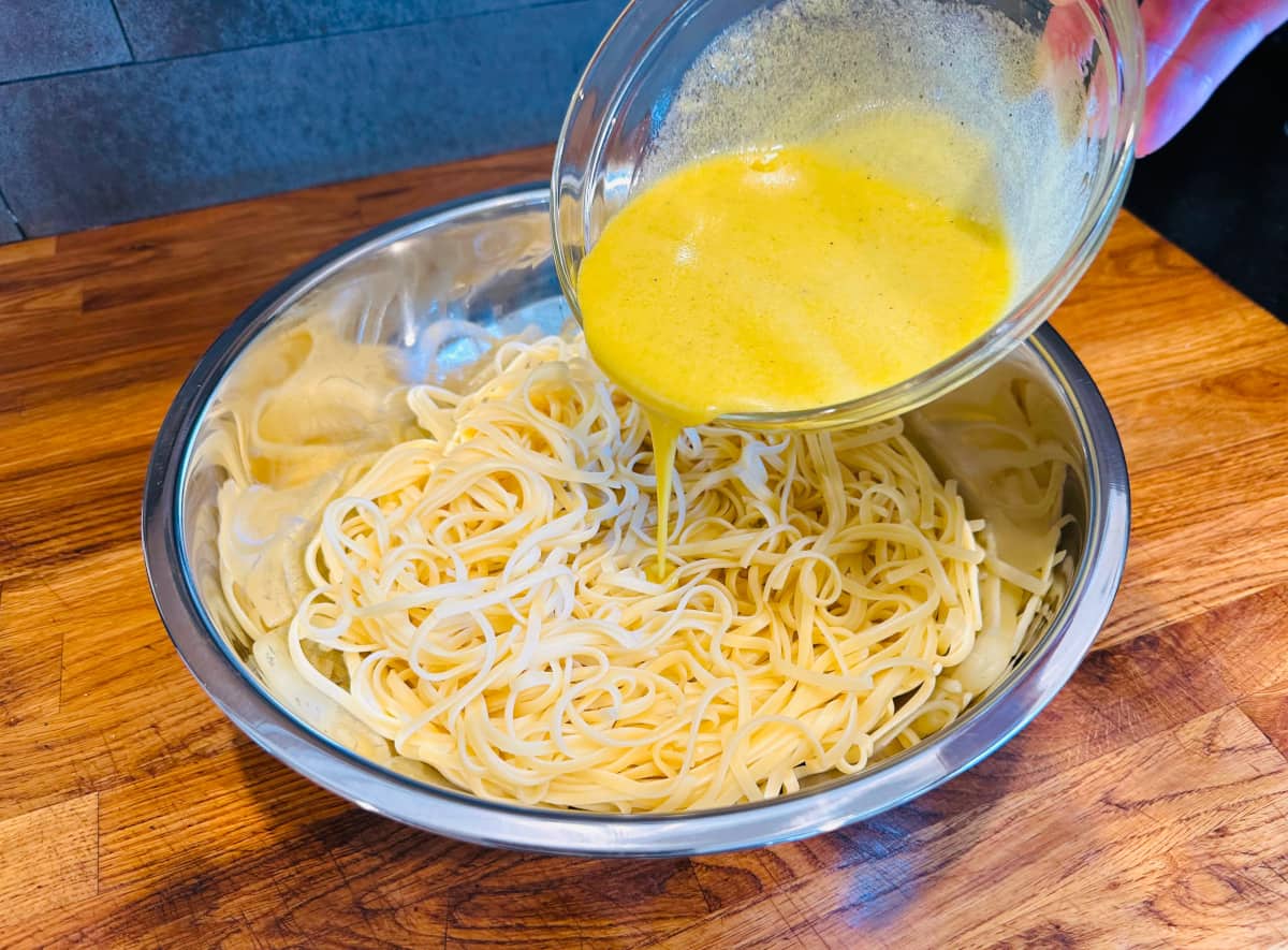 Yellow liquid being poured from a small glass bowl over cooked linguine in a large steel bowl.