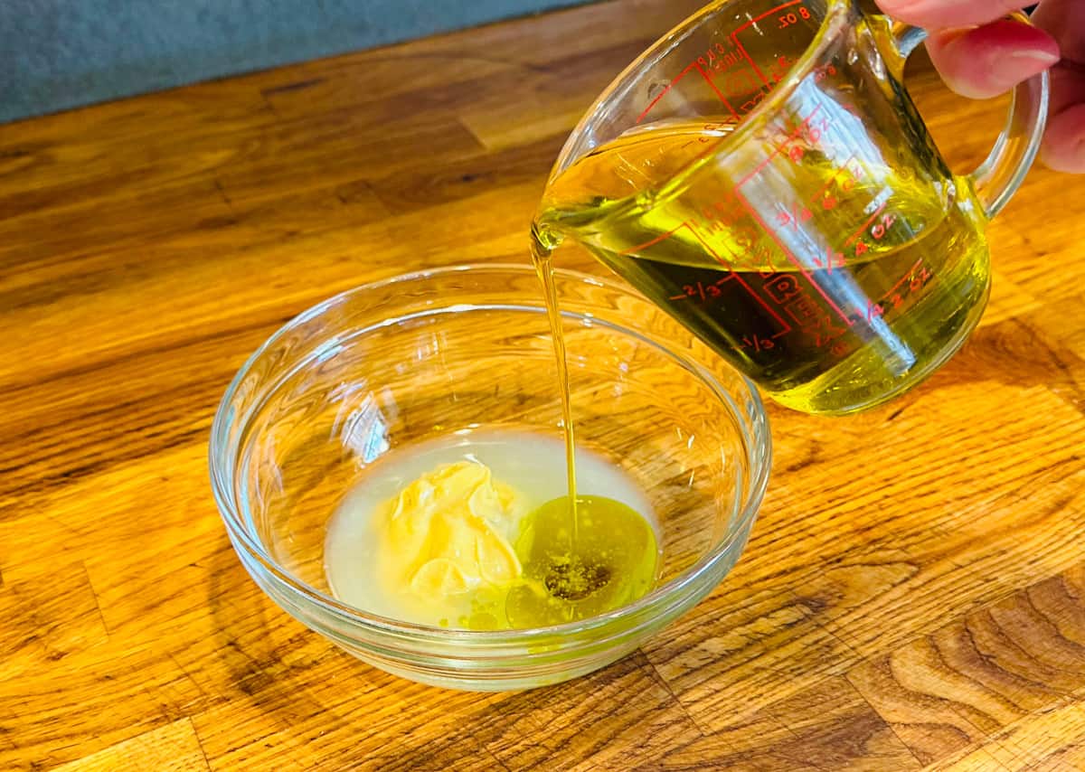 Olive oil being poured from a glass measuring cup into a small glass bowl containing lemon juice and a large dollop of dijon mustard.