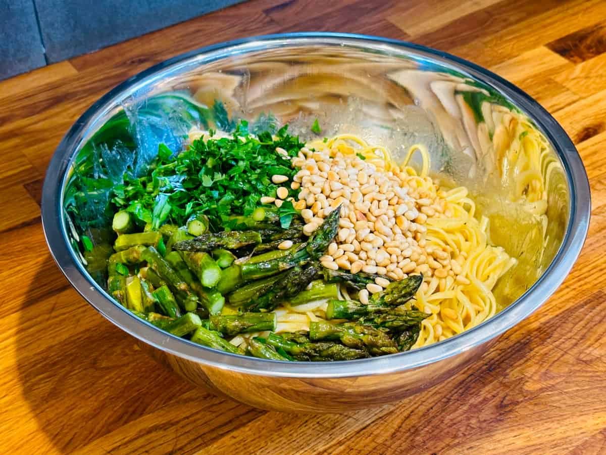 Pieces of roasted asparagus, chopped parsley, and pine nuts on top of cooked linguine in a large steel bowl.