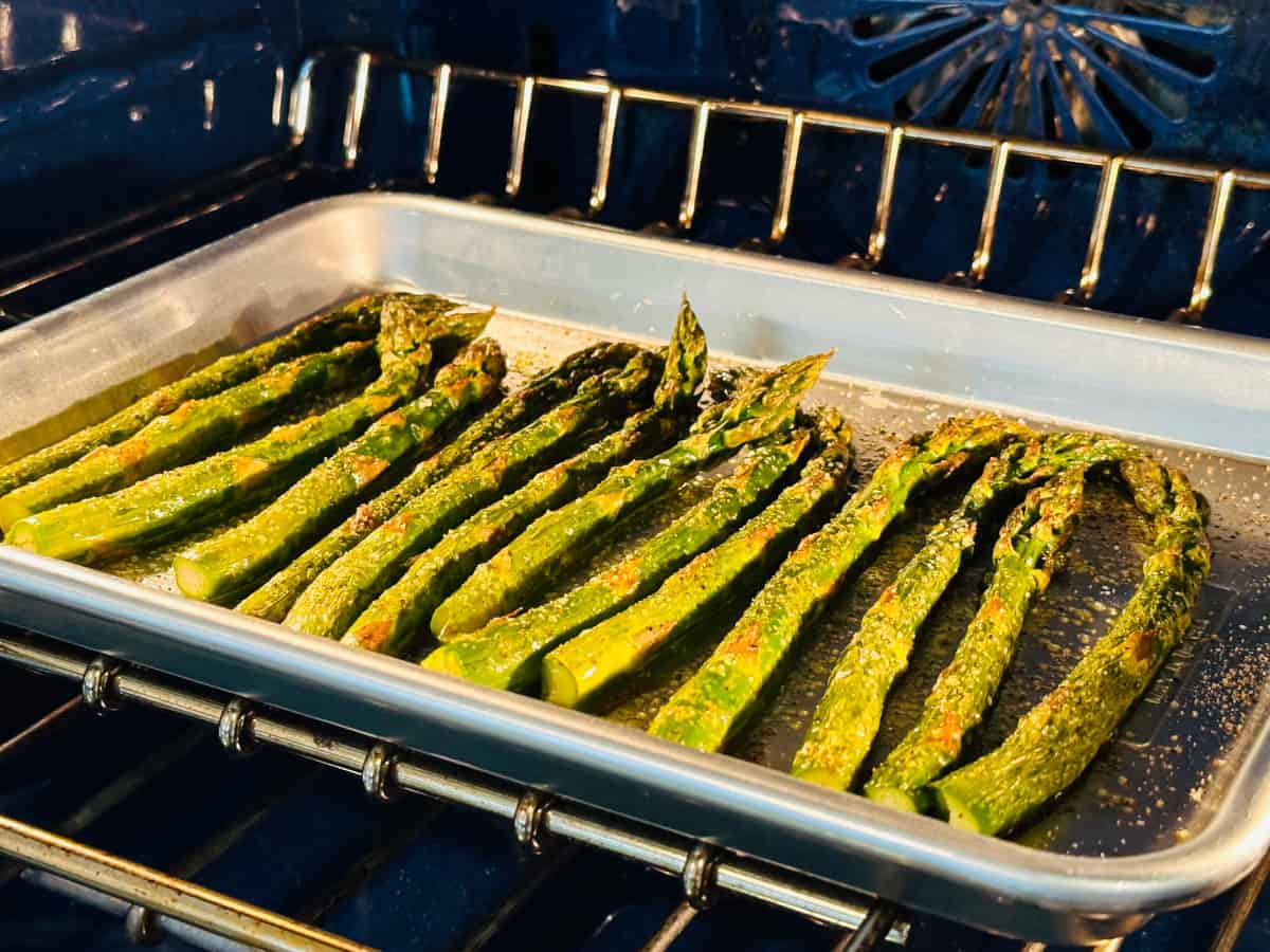 Asparagus spears on a small metal baking sheet roasting in a blue oven.
