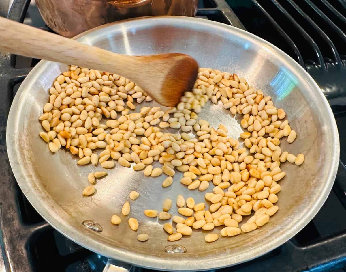 Pine nuts being stirred with a wooden spoon in a small saucepan on the stove.