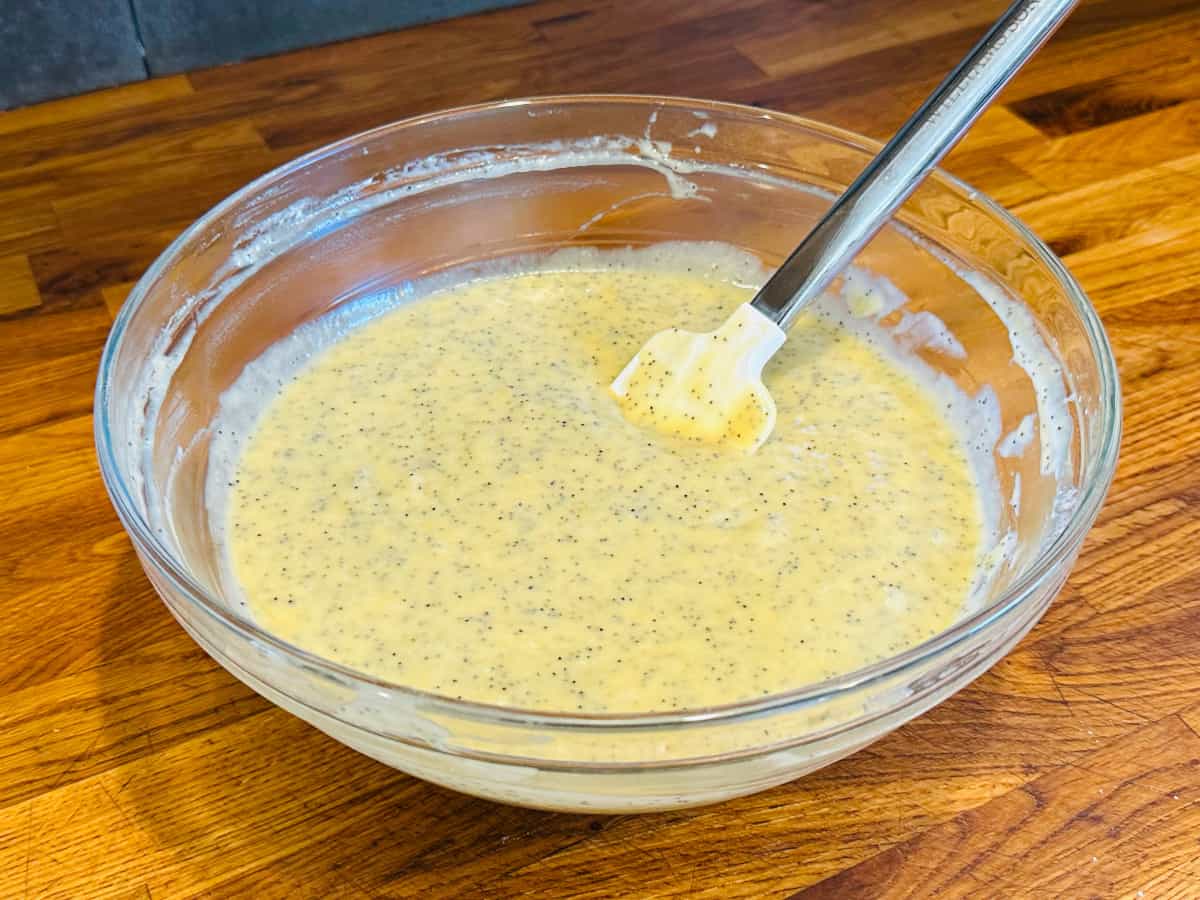 Pale yellow batter flecked with poppy seeds in a glass bowl with a white silicone spatula.