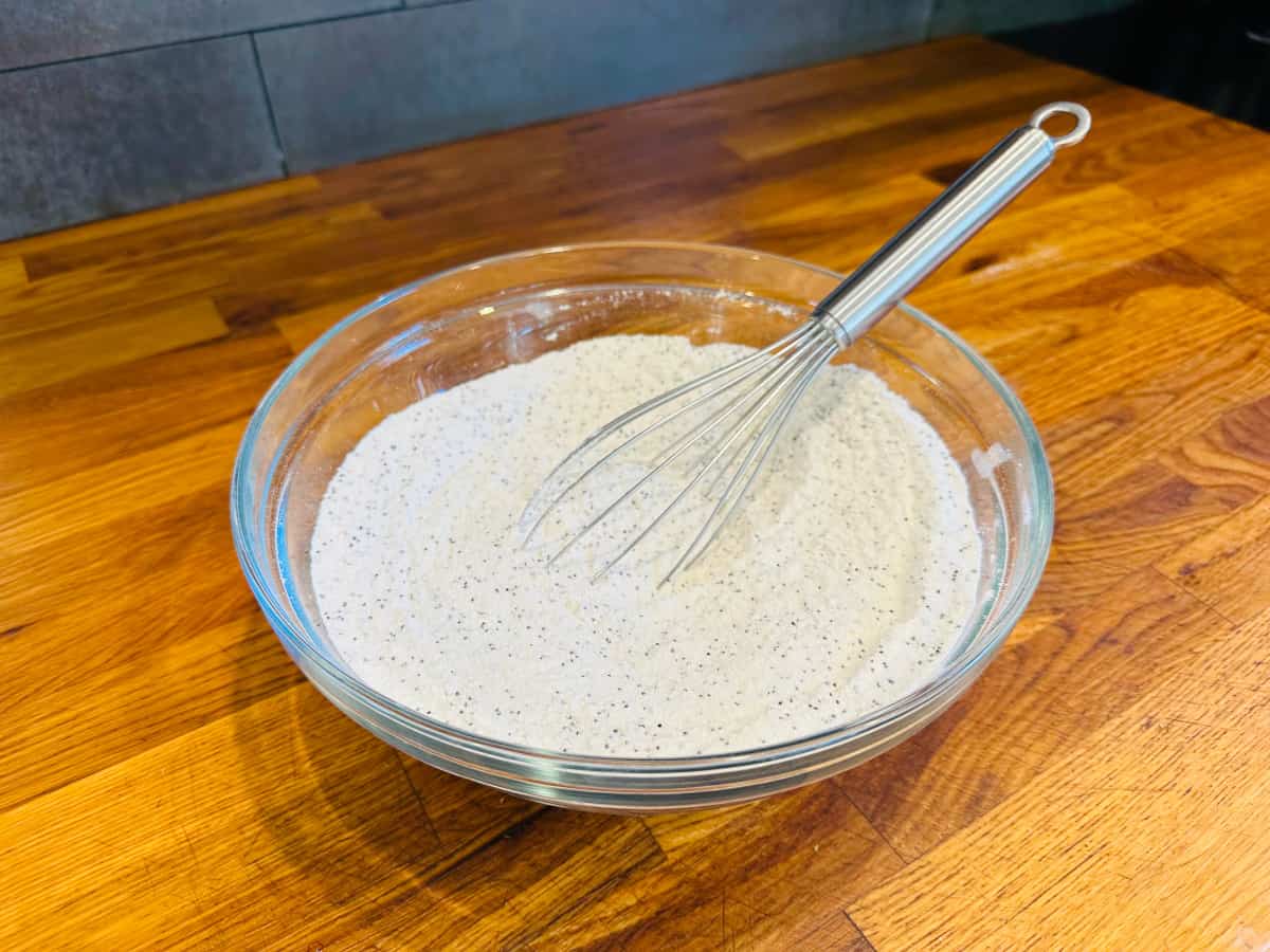 Dry ingredients flecked with poppy seeds in a glass bowl with a steel whisk.