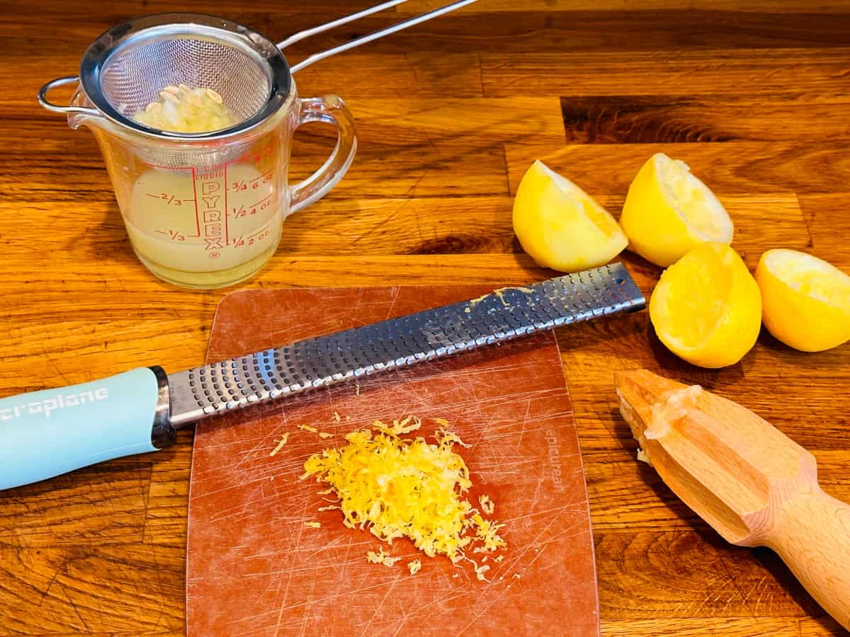 Lemon juice in a glass measuring cup with a strainer full of seeds on top next to a microplane, lemon zest, juiced lemon halves, and a wooden citrus reamer.