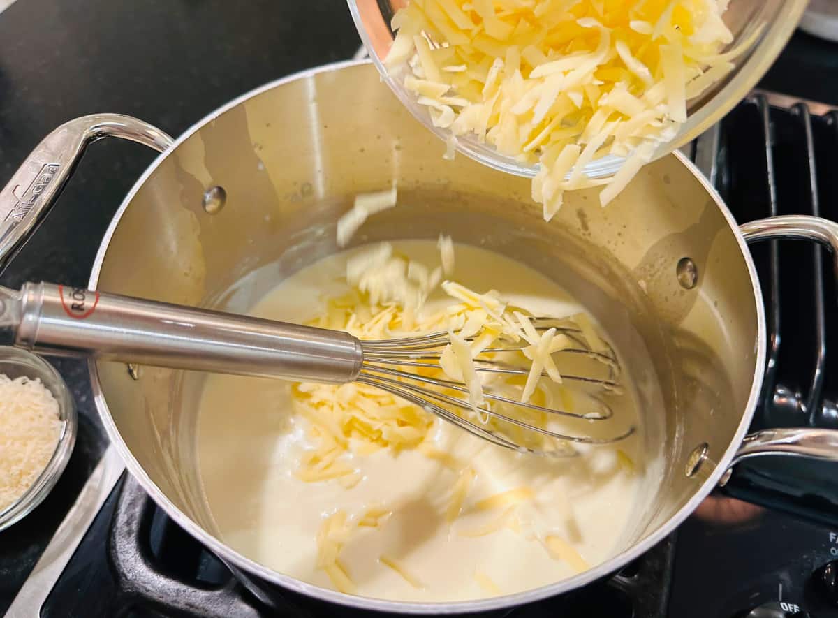 Shredded white cheese being sprinkled into cream colored sauce in a metal saucepan with a whisk.