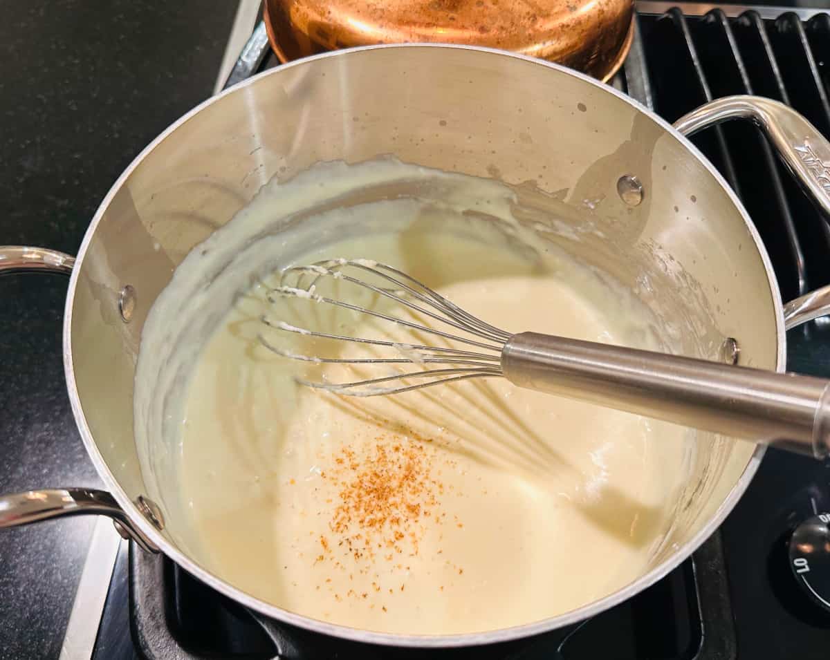 Cream colored sauce with a sprinkling of grated nutmeg on top in a metal saucepan with a whisk.