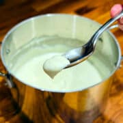 A dollop of cream colored sauce on the end of a spoon over a metal saucepan containing the same sauce.