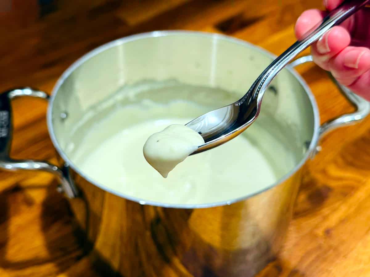 A dollop of cream colored sauce on the end of a spoon over a metal saucepan containing the same sauce.