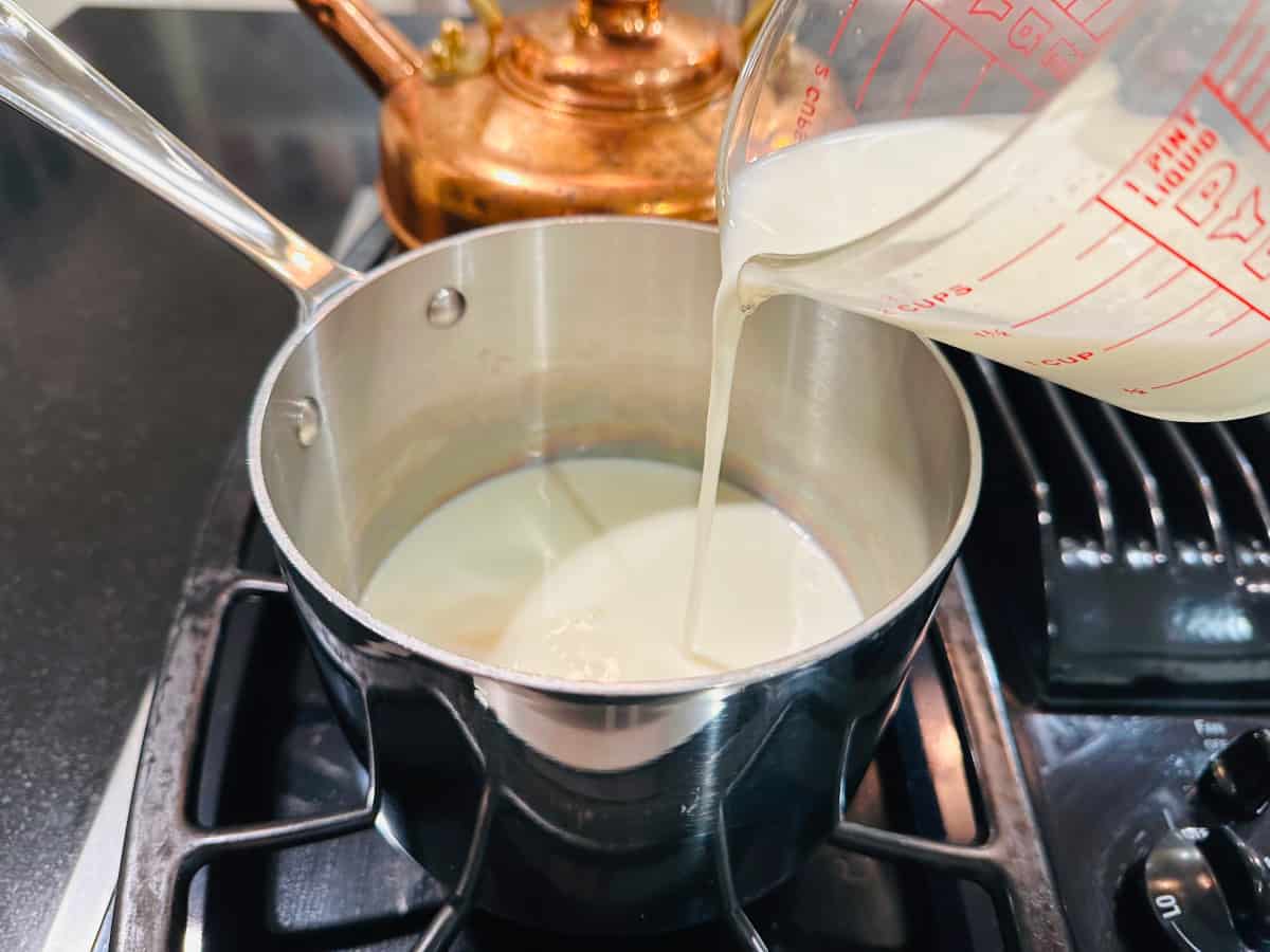 Milk being poured from a glass measuring cup into a small steel saucepan on the stove.