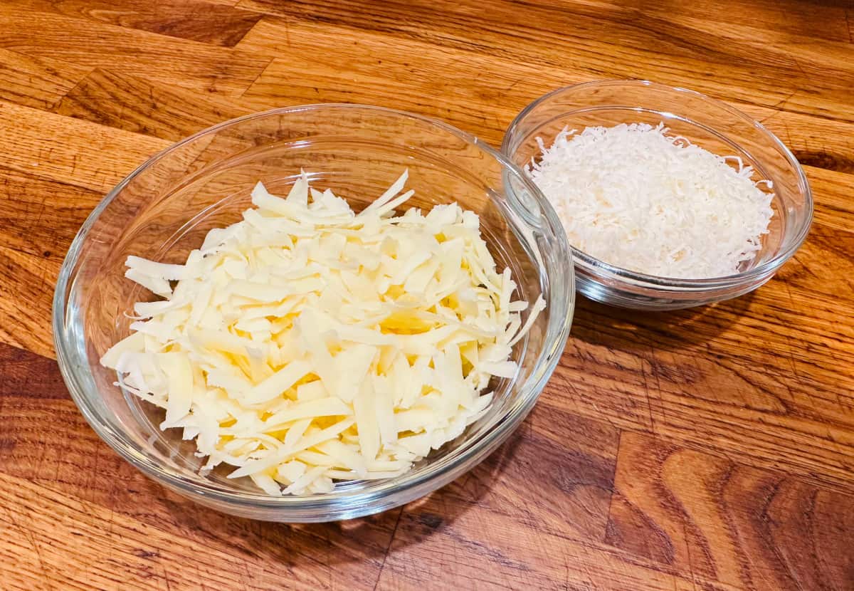 Shredded Gruyere in a medium glass bowl next to finely shredded Parmigiano Reggiano in a small glass bowl.