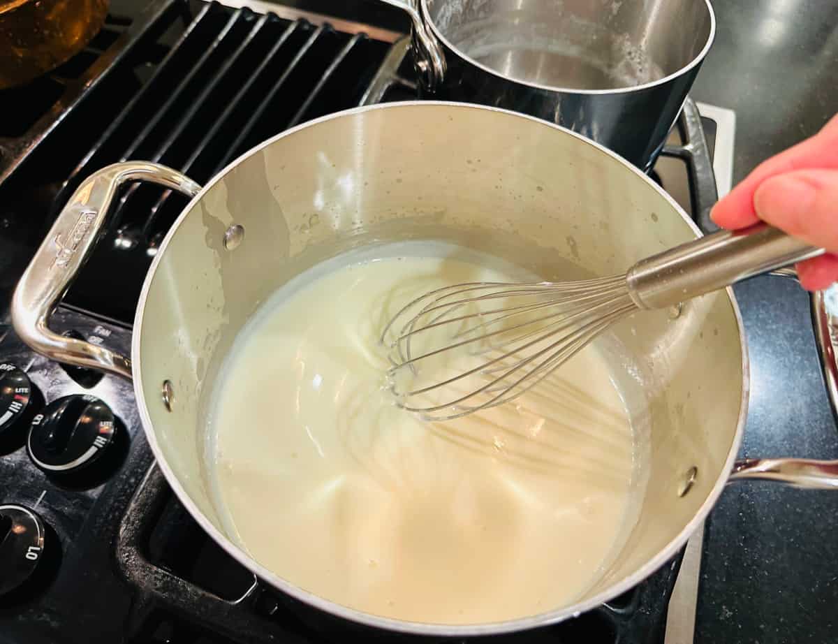 Cream colored sauce being whisked in a metal saucepan on the stove.