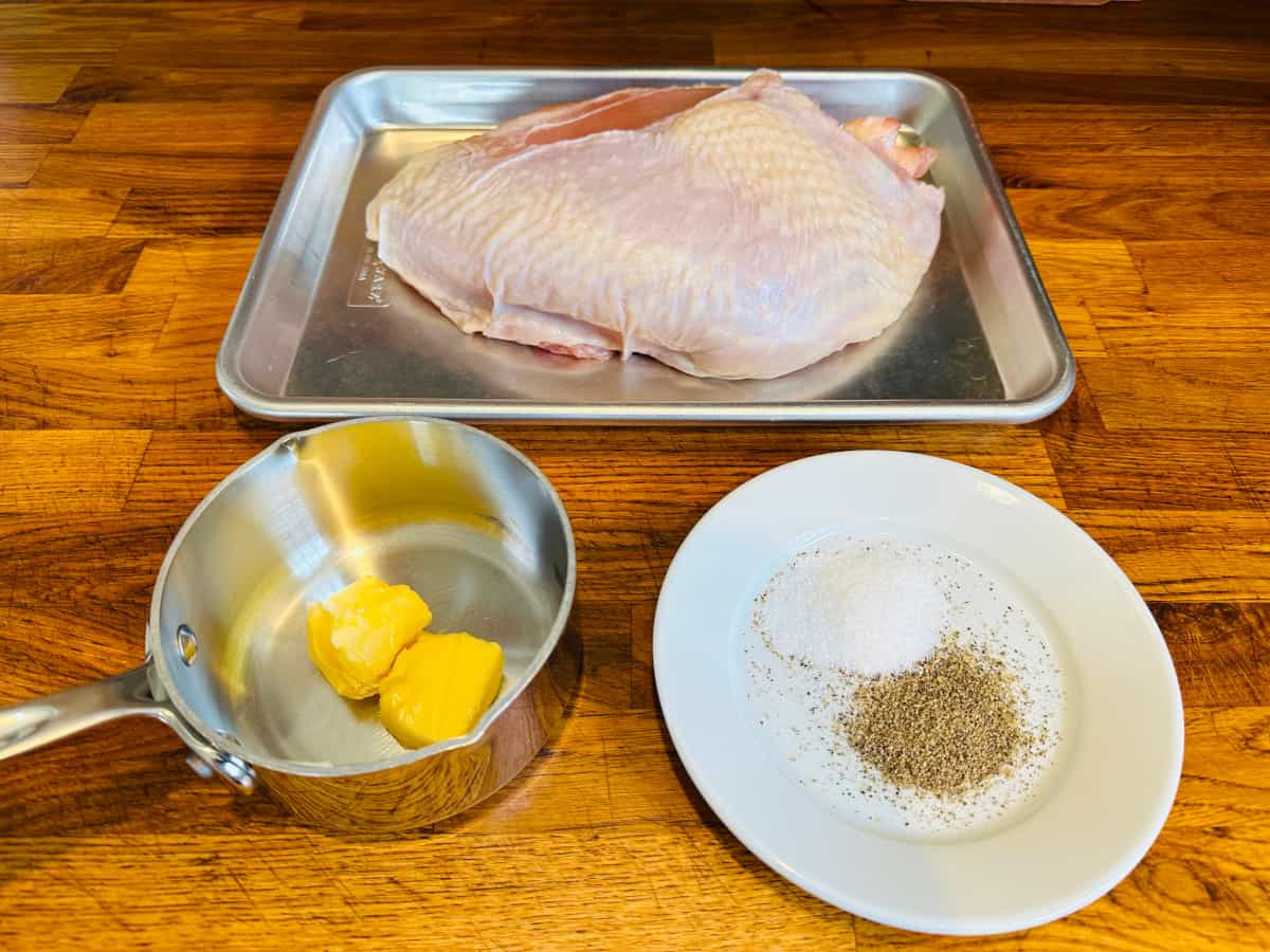 Raw turkey breast on a metal half baking sheet behind a small steel saucepan with two cubes of butter and piles of salt and pepper on a small white plate.