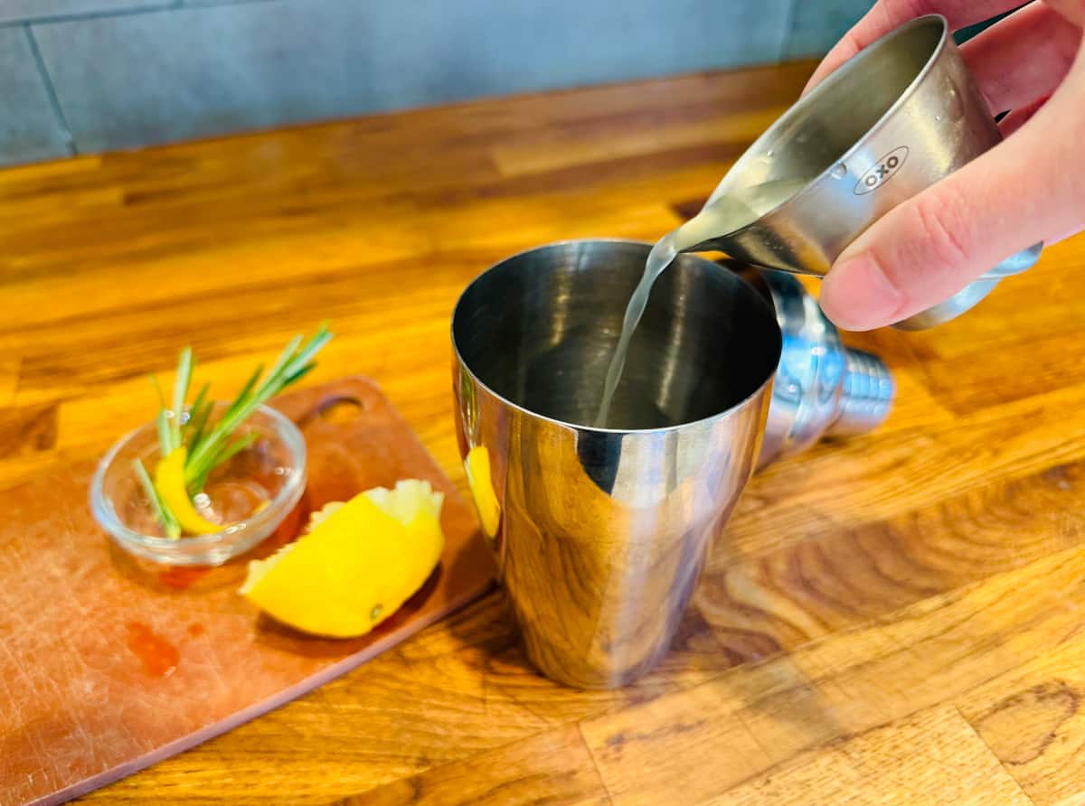 Pale yellow liquid being poured from a steel measuring jigger into a cocktail shaker next to a squeezed half lemon and a small glass bowl containing a lemon twist and a couple sprigs of rosemary.
