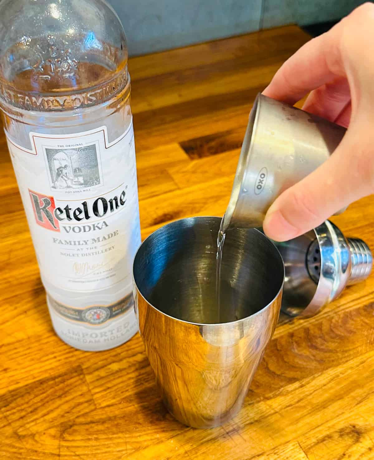 Clear liquid being poured from a steel measuring jigger into a cocktail shaker next to a bottle of vodka.