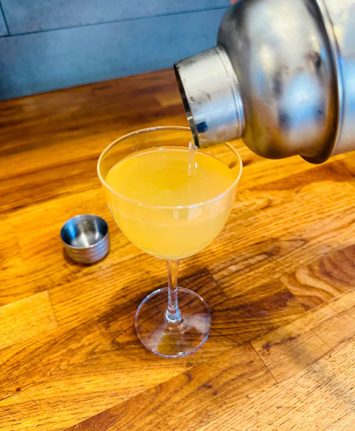 Pale yellow liquid being poured from a cocktail shaker into a coupe glass.