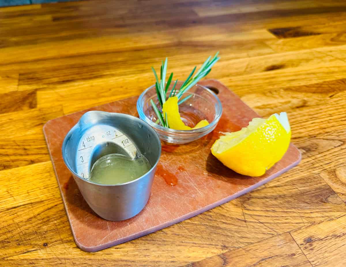 Pale yellow liquid in a steel measuring jigger next to a squeezed half lemon and a small bowl containing a lemon twist and a couple sprigs of rosemary.