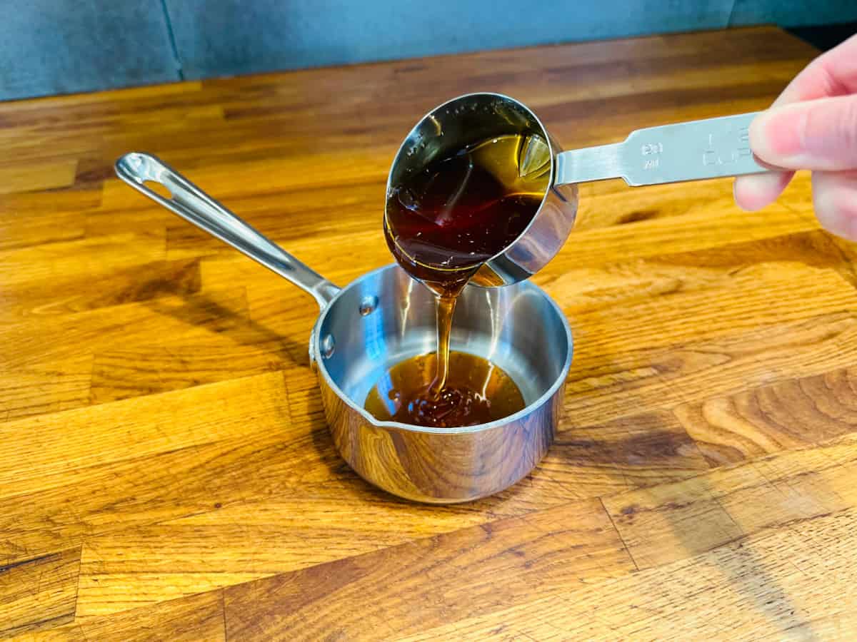 Golden brown honey being poured from a metal measuring cup into a small steel saucepan.