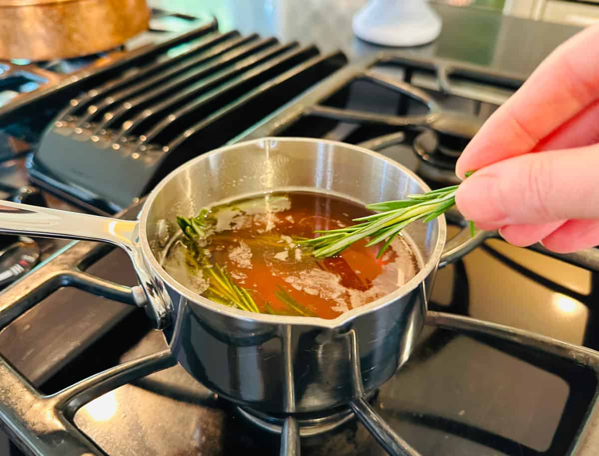 Sprigs of rosemary being added to golden brown liquid in a small steel saucepan on the stove.