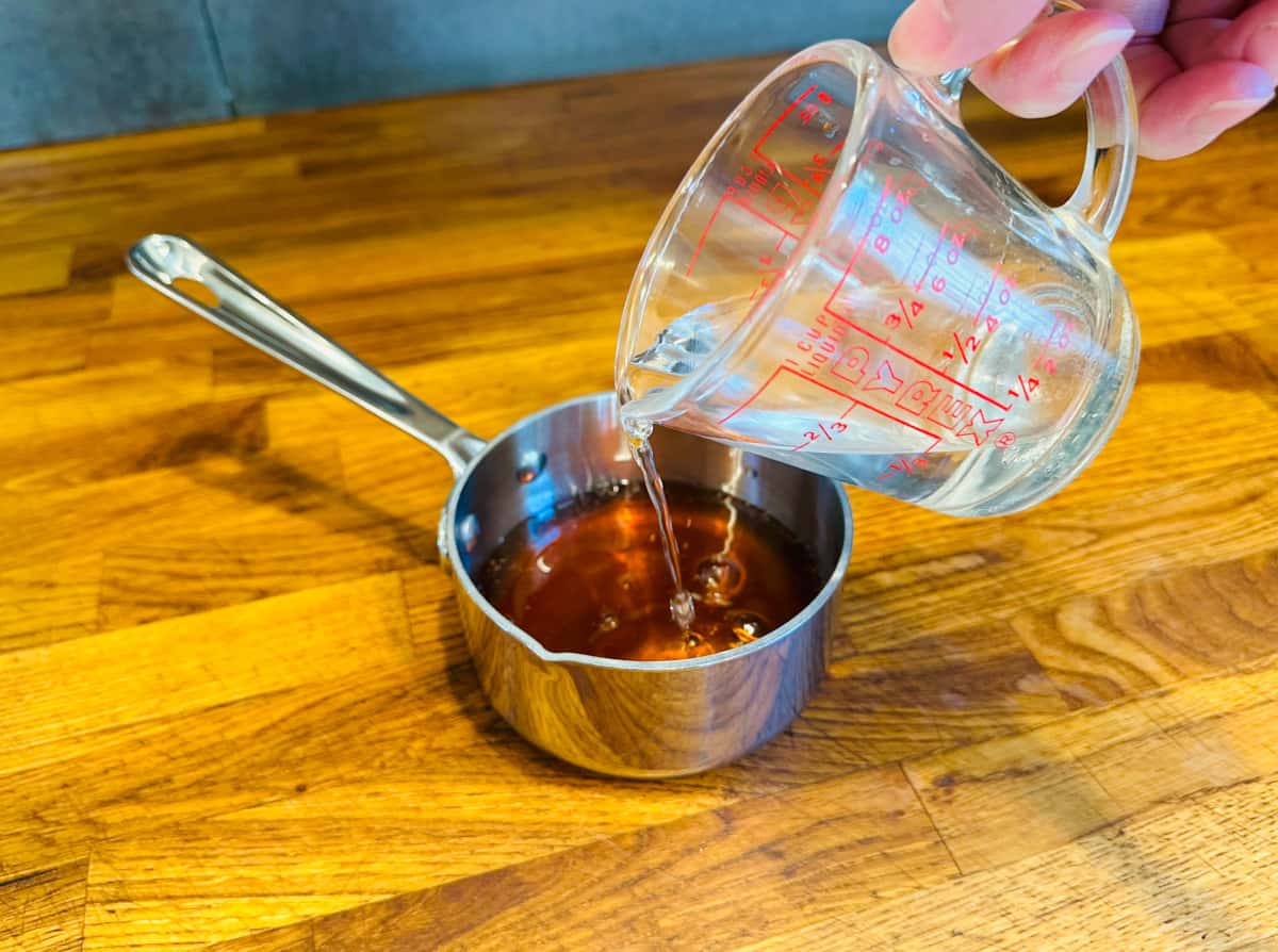 Water being poured from a glass measuring cup into golden brown honey in a small steel saucepan.