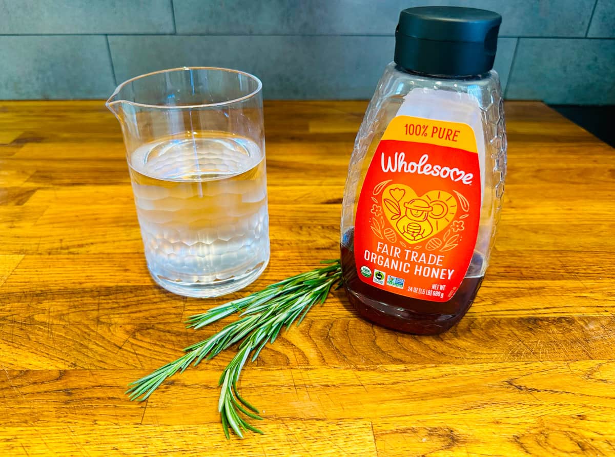 A small glass pitcher of water, a few sprigs of rosemary, and a bottle of honey with an orange label.