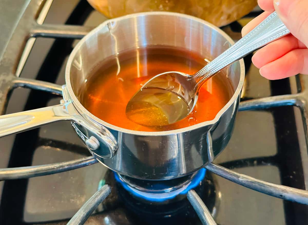 Golden brown honey and water being stirred with a steel spoon in a small steel saucepan over a low flame on the stove.
