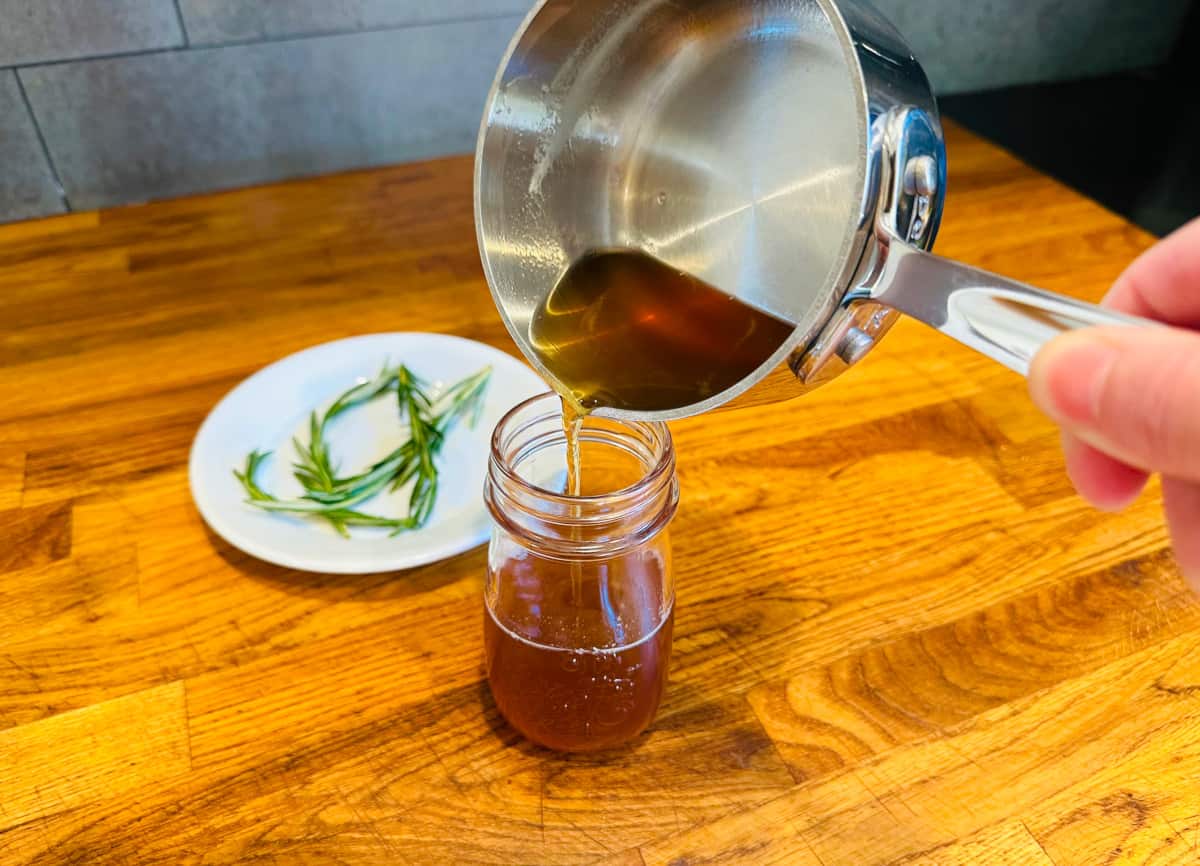 Rosemary simple syrup being poured from a small steel saucepan into a small bottle next to rosemary sprigs on a white plate.