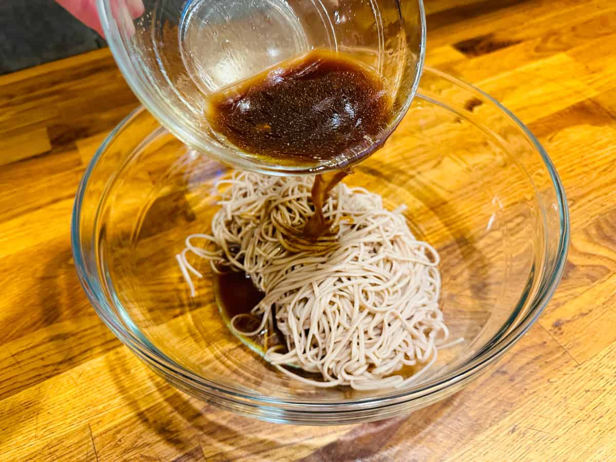 Dressing being poured over cooked soba noodles in a glass bowl.