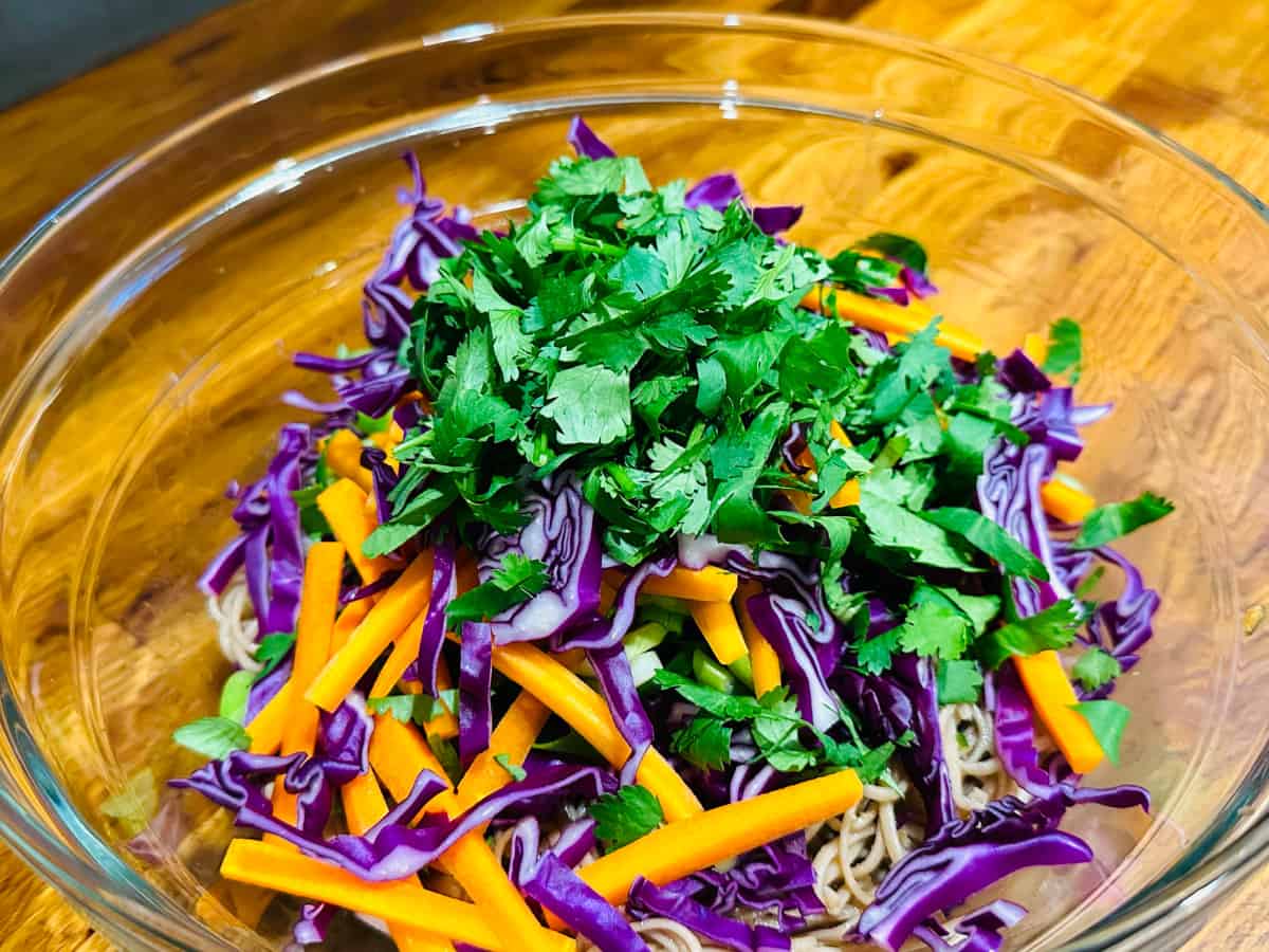 A pile of chopped cilantro, carrots, and red cabbage on top of soba noodles in a glass bowl.