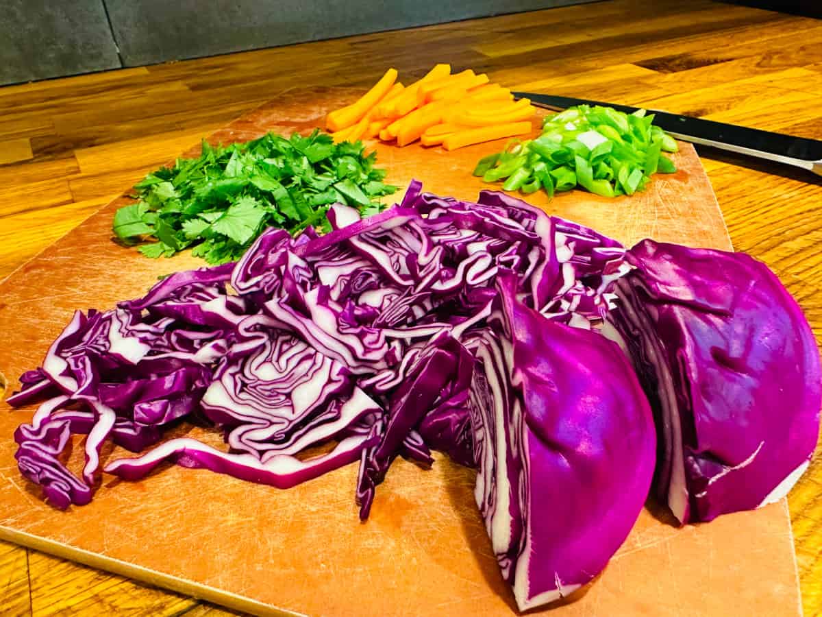 Thinly sliced red cabbage, chopped cilantro, chopped scallions, and matchstick carrots on a cutting board with a knife.