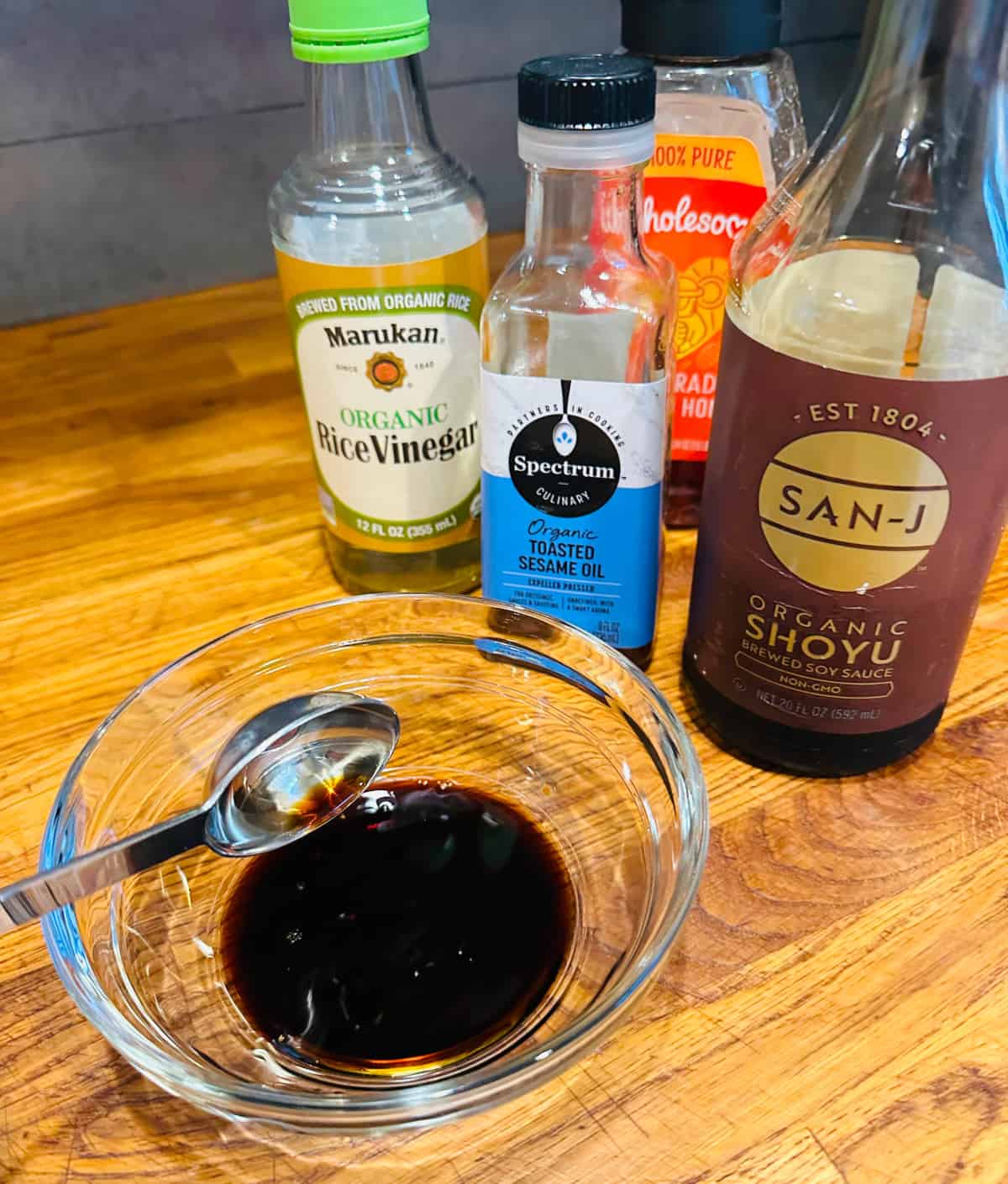 Shoyu being poured from a steel tablespoon into a small glass bowl next to bottles of shoyu, rice vinegar, toasted sesame oil, and honey.