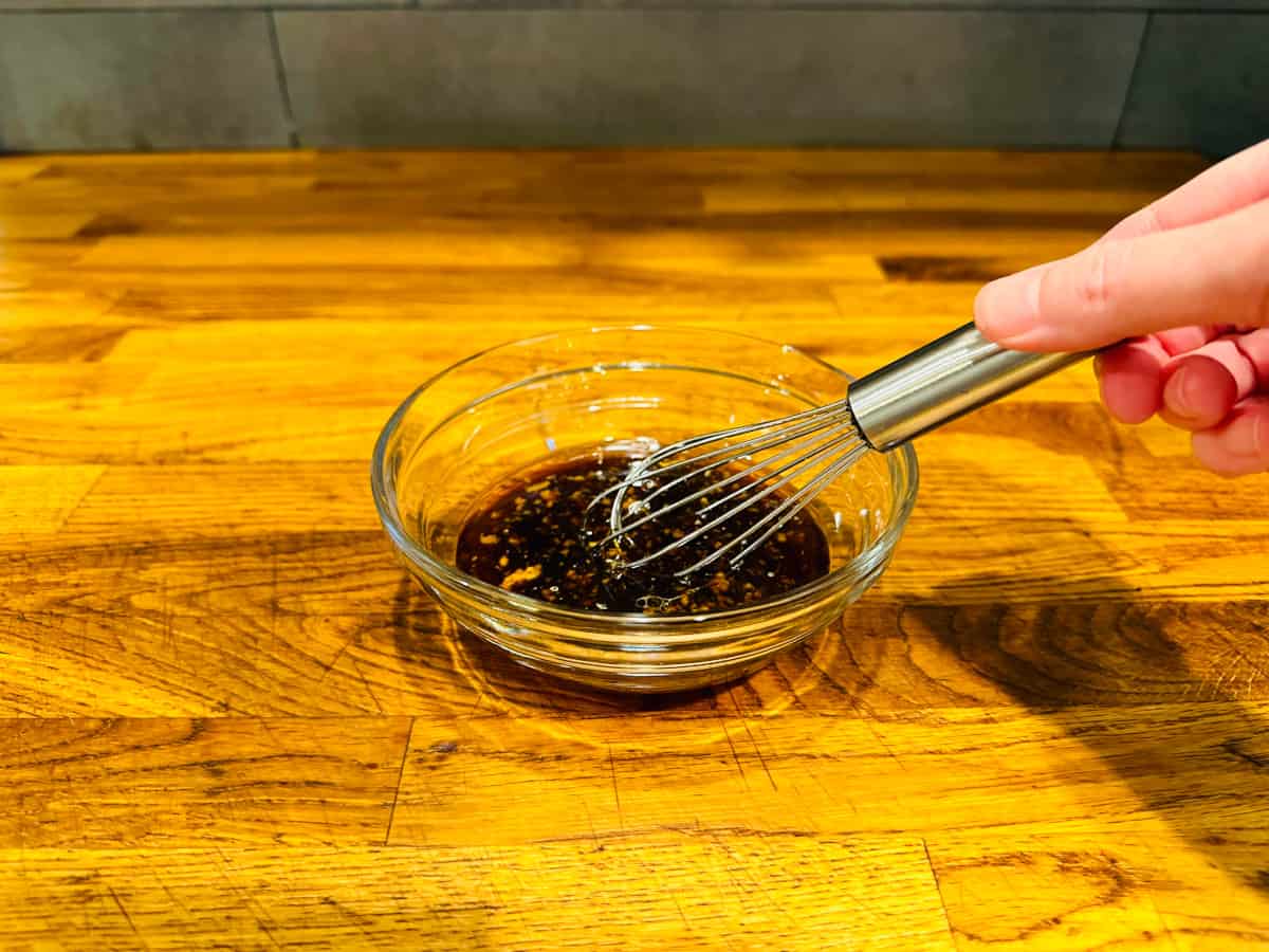 Dark brown liquid flecked with grated ginger being whisked in a small glass bowl.