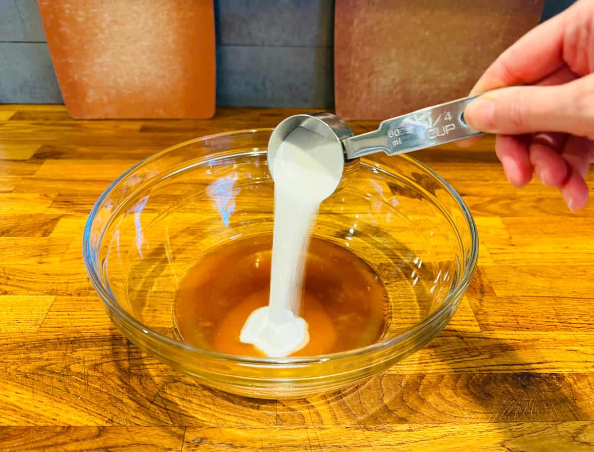 Sugar being poured from a small steel measuring cup into a glass bowl containing light brown liquid.