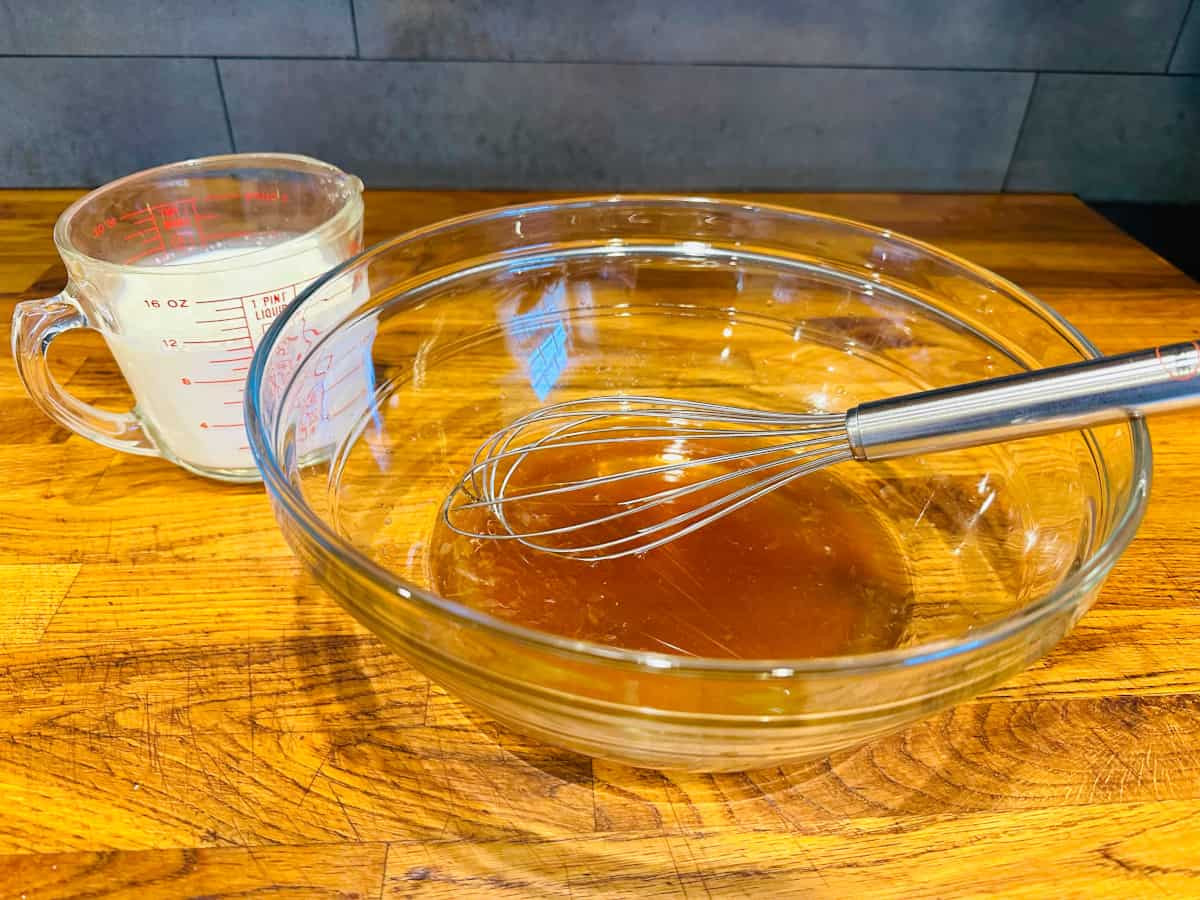 Glass measuring cup of cream next to a glass bowl containing light brown liquid and a metal whisk.