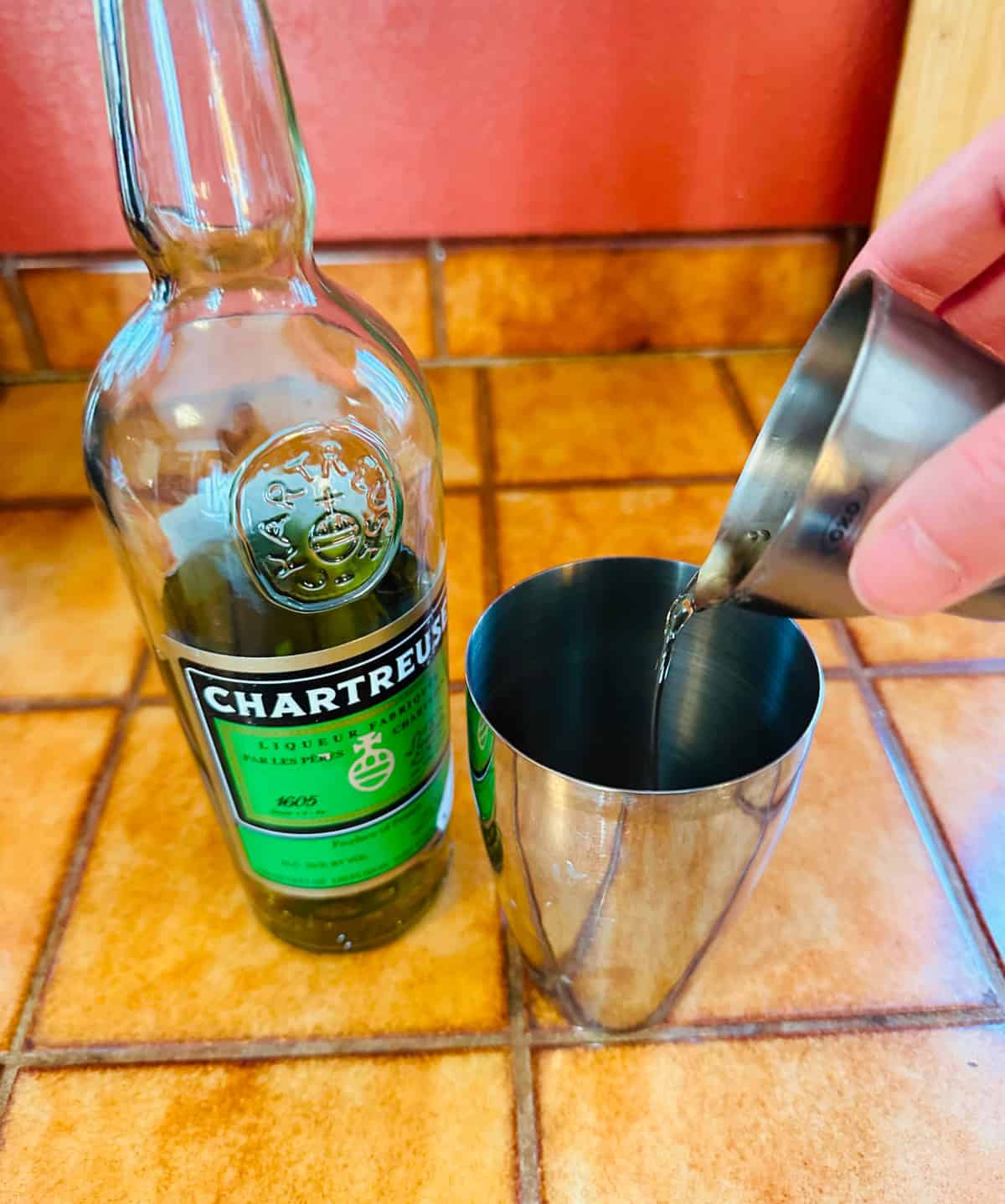 Pale green liquid being poured from a steel measuring jigger into a cocktail shaker next to a bottle of chartreuse.