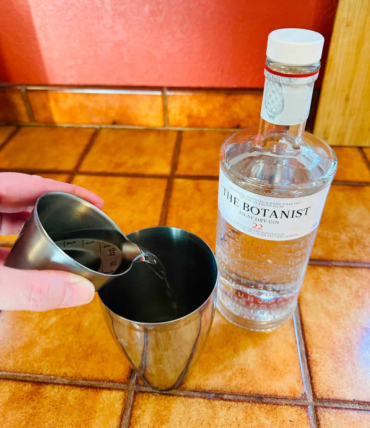 Clear liquid being poured from a steel measuring jigger into a cocktail shaker next to a bottle of gin.