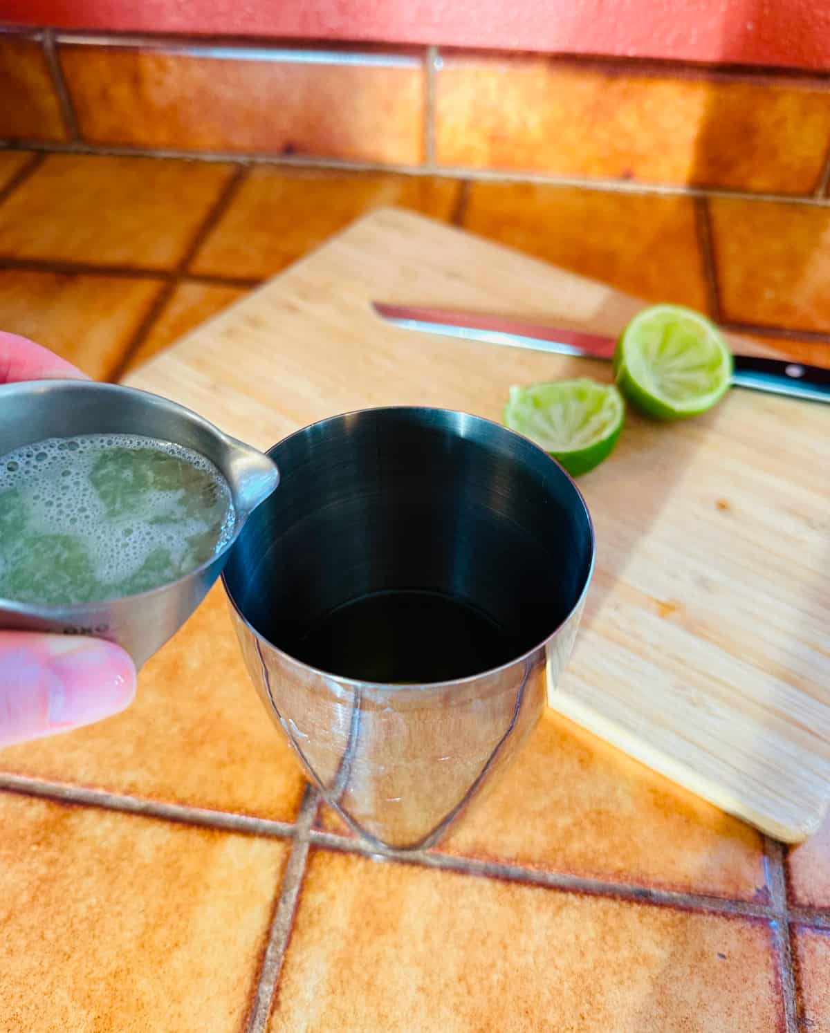 Pale green juice being poured from a steel measuring jigger into a cocktail shaker next to two halves of lime on a cutting board with a knife.