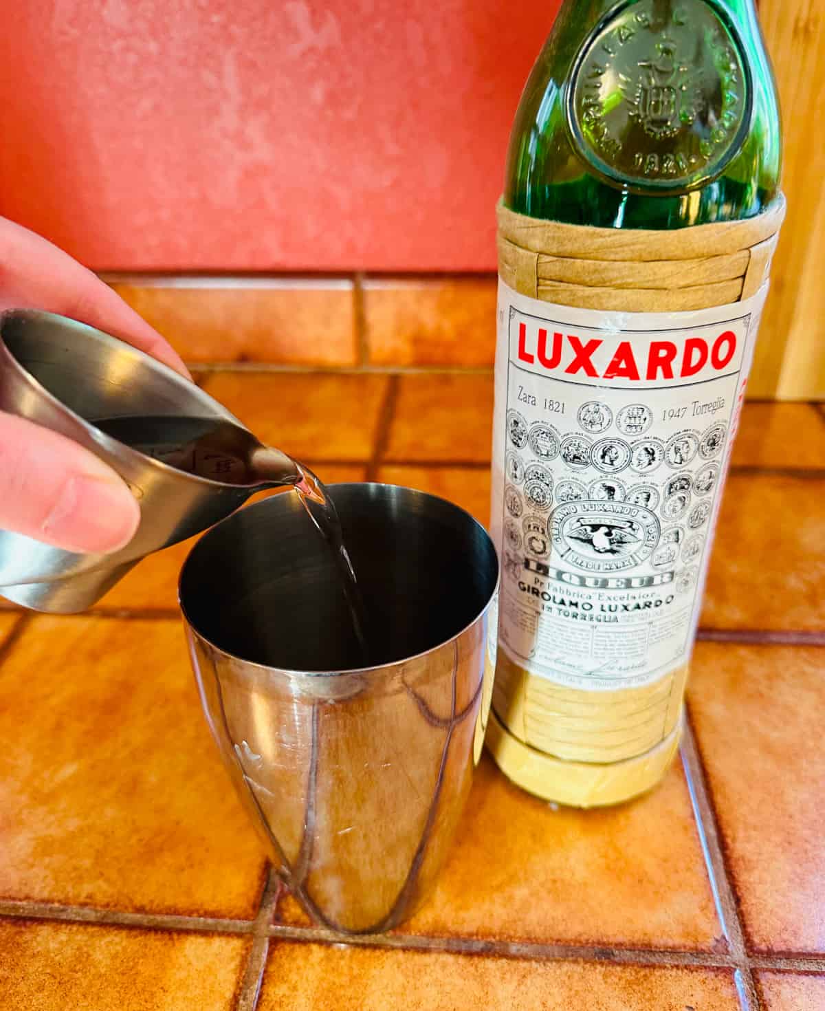 Pale yellow liquid being poured from a steel measuring jigger into a cocktail shaker next to a bottle of luxardo.