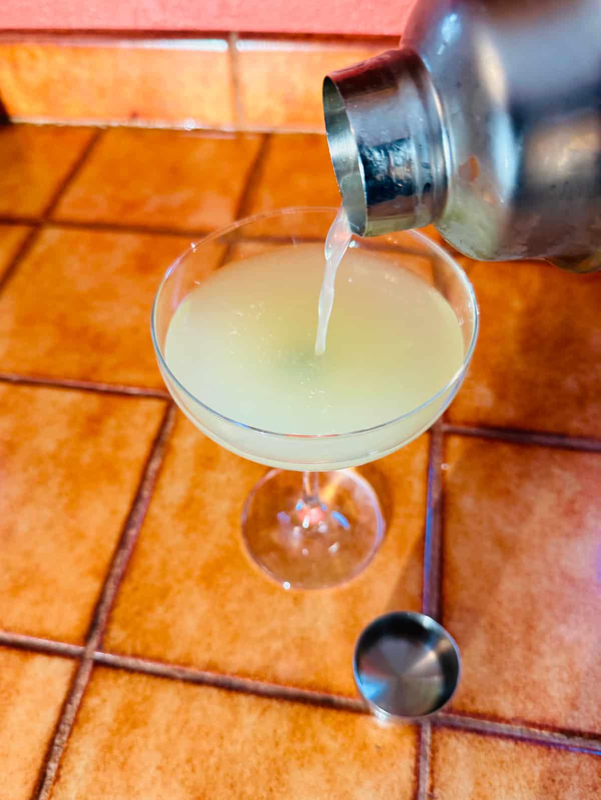 Pale green liquid being poured out of a cocktail shaker into a coupe glass.